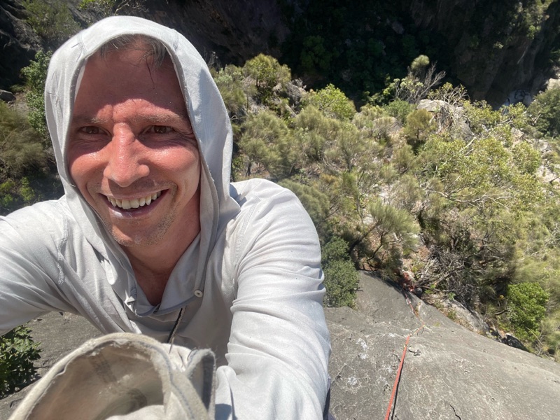 Duncan in a grey hood, smiling, high on a rock face with bush and a rope below.